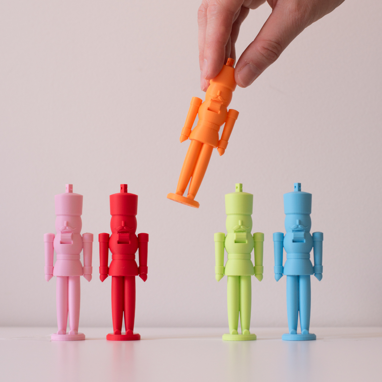 Five colorful nutcracker figures in pastel tones (pink, red, orange, green, and blue) on a white background. Hand holding the orange figure.