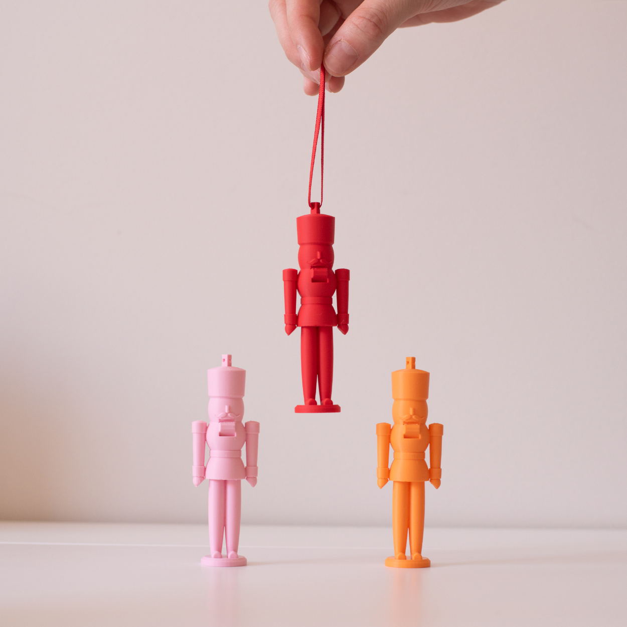 Three pastel nutcracker figures in pink, red, and orange on a light background, with one red figure being held by a ribbon.