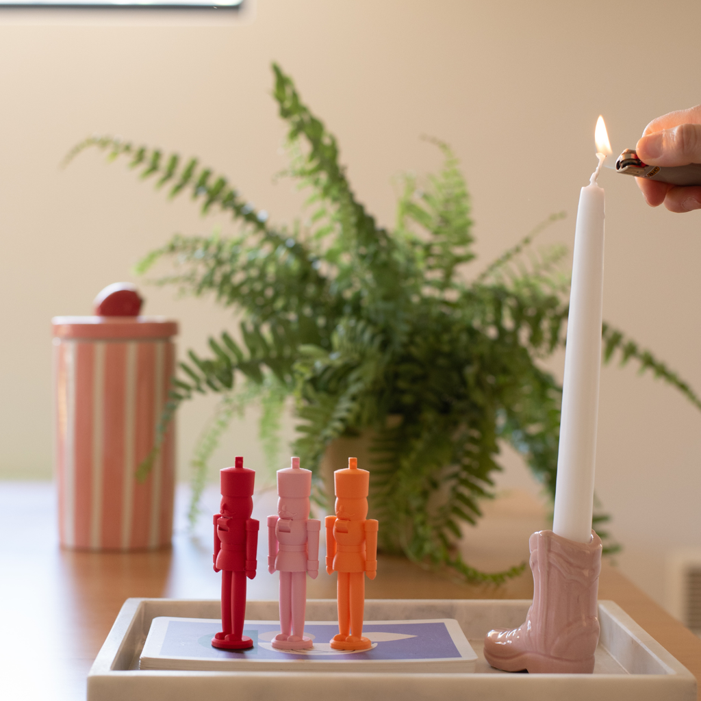 Three pastel nutcracker figures in red, pink, and orange beside a white candle in a pink boot-shaped holder, with a plant in the background.